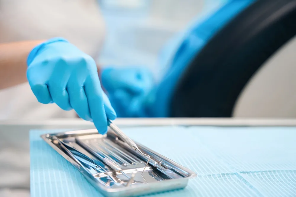 Blue-gloved hand selecting a dental instrument from a metal tray
